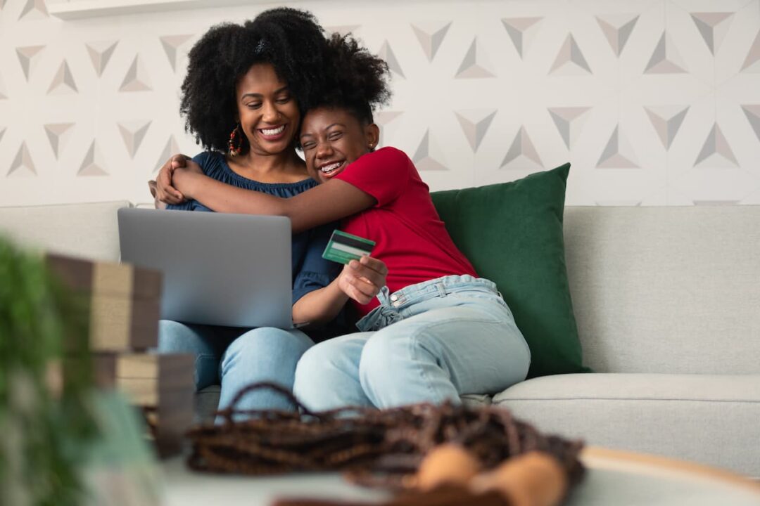 Mãe e filha negras se abraçando e sorrindo. estão sentadas em um sofá branco com um notebook no colo segundo um cartão