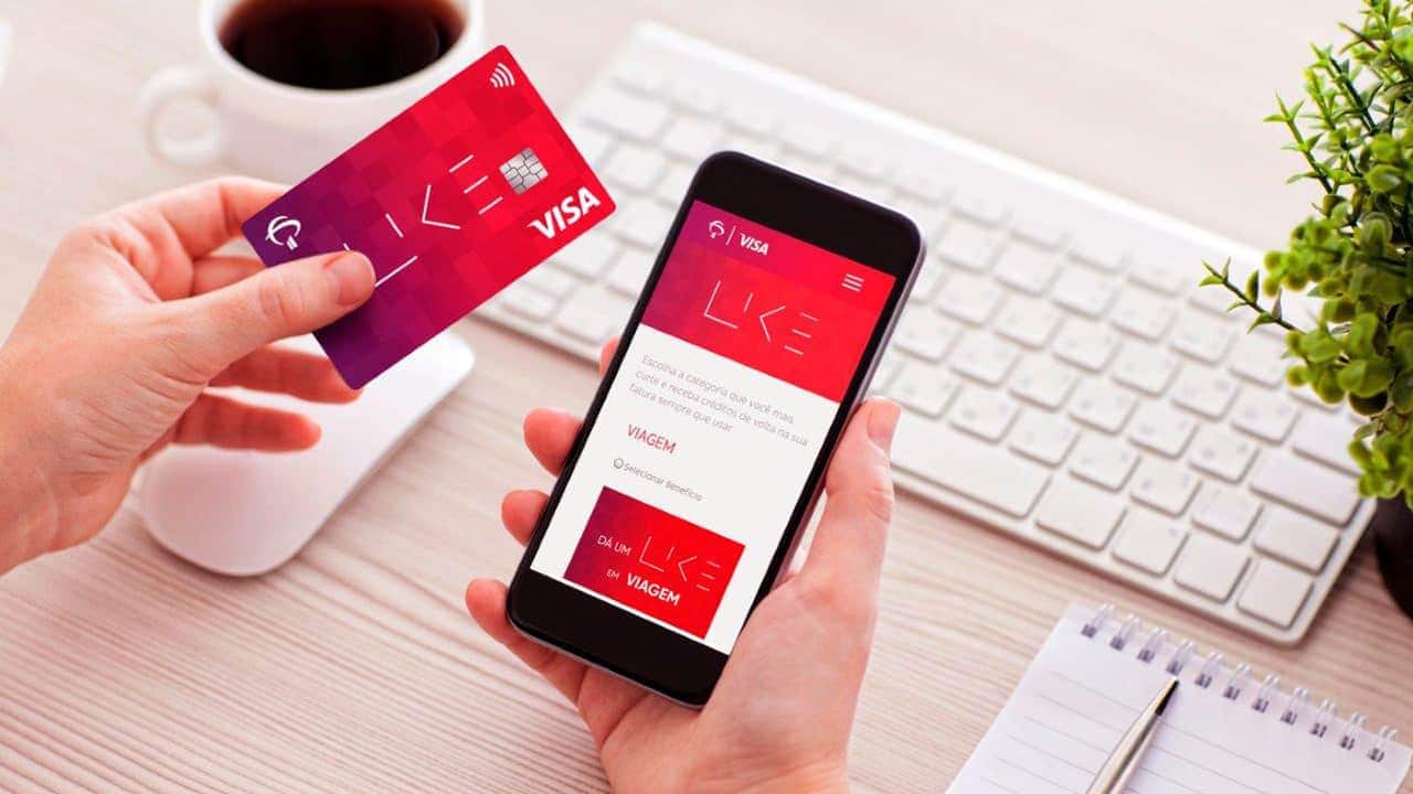 Imagem de pessoa segurando seu cartão Like Bradesco numa mão e na outra, seu celular com o app do Bradesco aberto