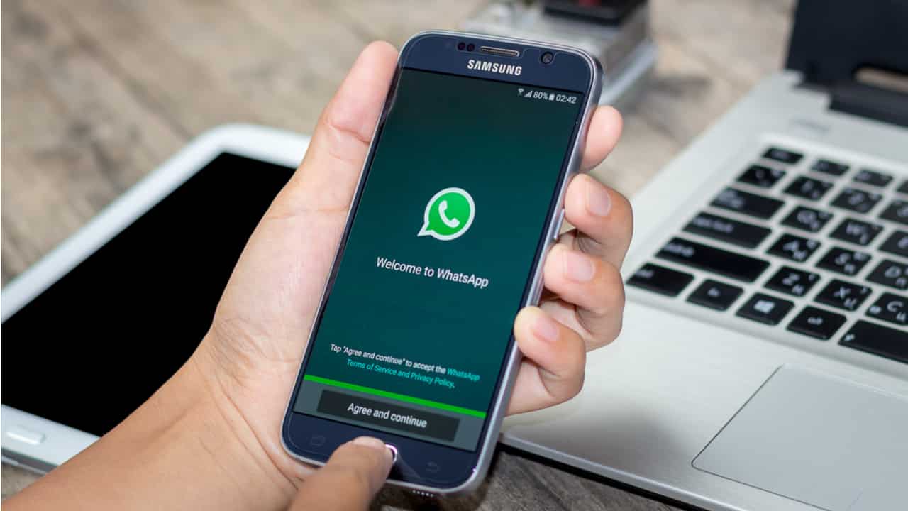 Imagem de um celular com o WhatsApp aparecendo na tela. Uma mão de pessoa branca segura o celular. Ao fundo, sobre uma mesa, há um tablet desligado e um notebook