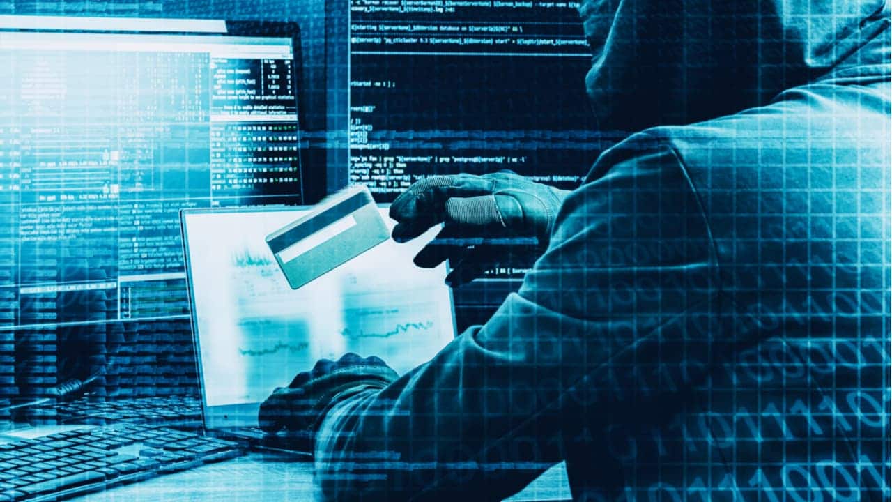 pessoa encapuzada com cartão de crédito em mãos em frente ao computador