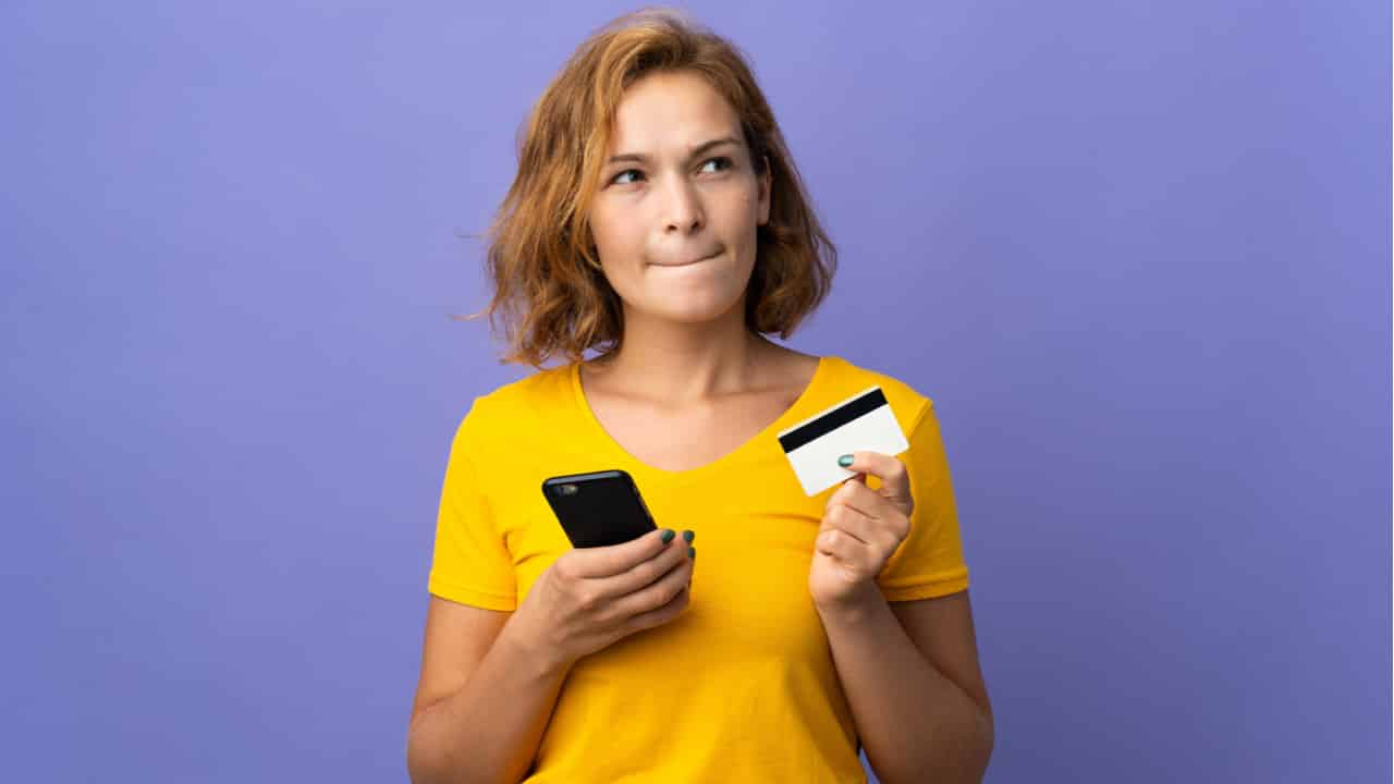 Imagem de mulher usando camisa amarela e segurando seu celular e um cartão, um em cada mão