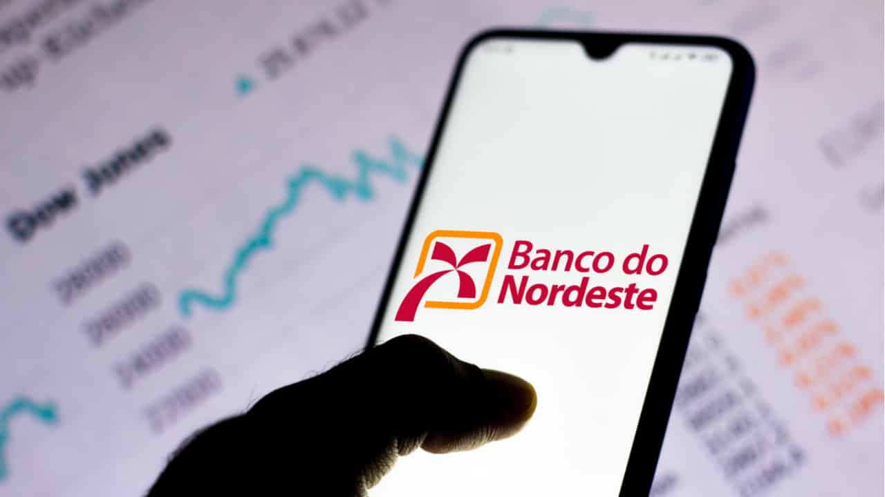 Celular sendo segurado por uma mão e com tela inicial destacando o logo do Banco do Nordeste. Ao fundo dados estatísticos