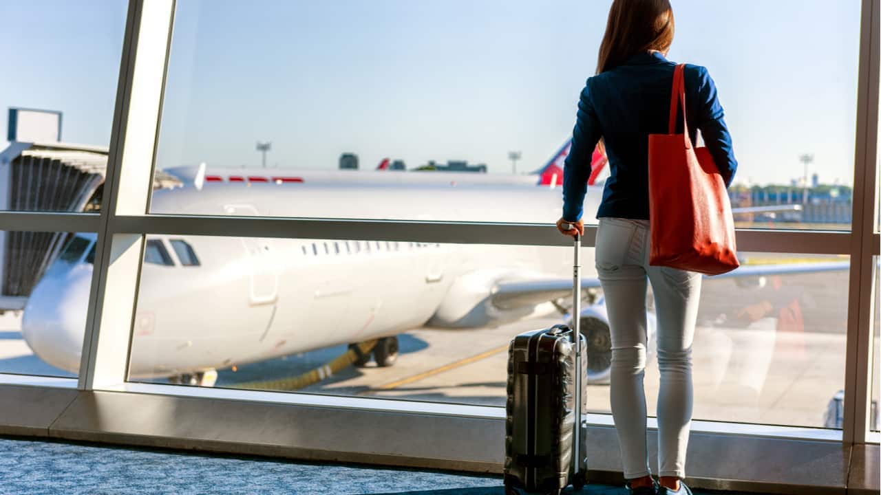 Mulher em saguão de aeroporto, de costas, com bagagem de mão, observando uma aeronave na pista de pouso