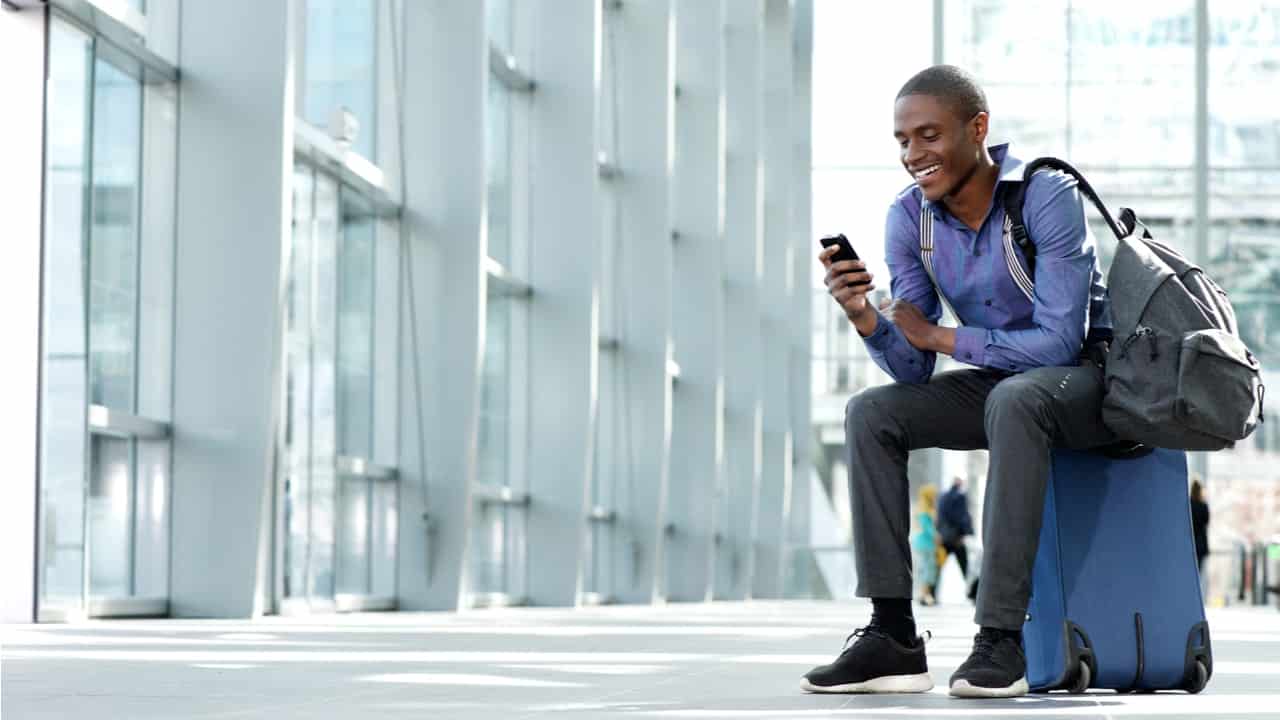 homem sorri enquanto olha para celular segurando uma mochila de viagem, sentado em cima de uma mala, no aeroporto
