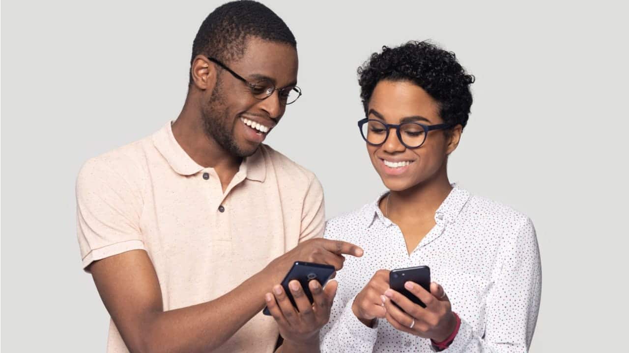 Dois jovens negros sorrindo olhando pros celulares um do outro após receberem quantia de banco por meio do Indique e Ganhe