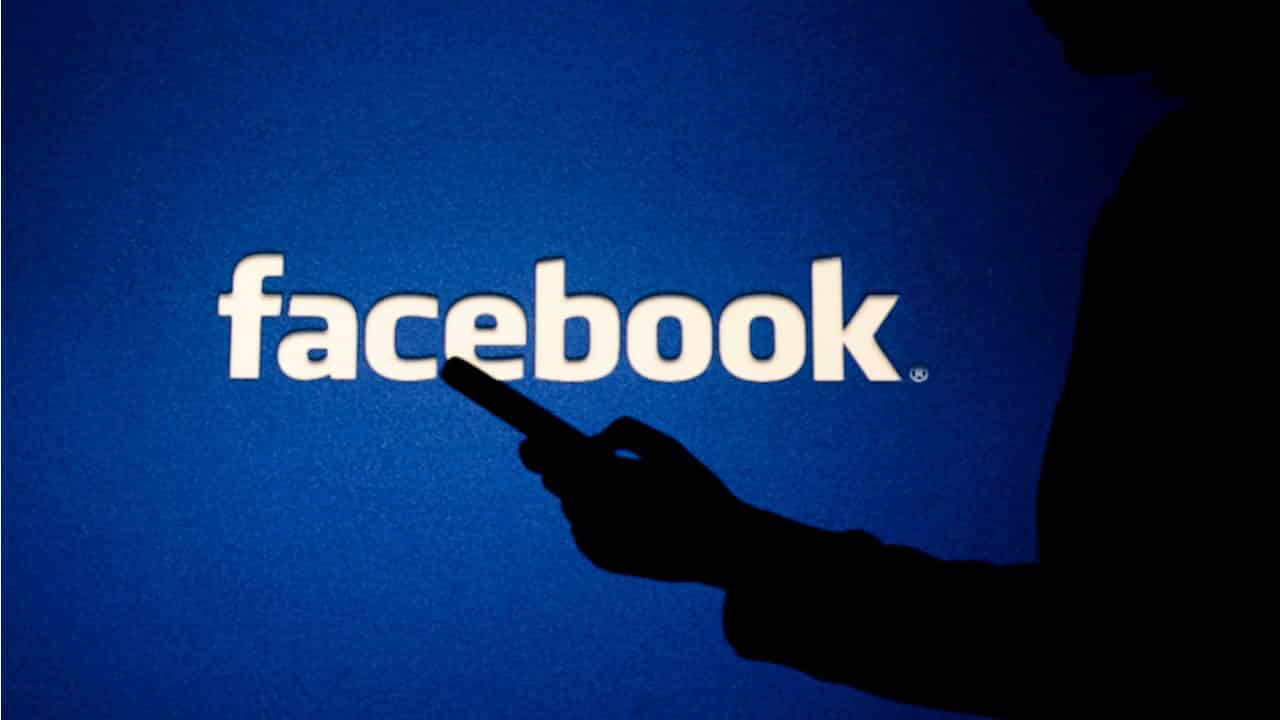 Sombra de pessoa segurando celular em frente a um telão da logo do facebook.