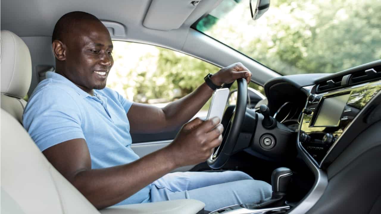 Motorista dentro de um carro olhando para o celular em uma das mãos e sorrindo enquanto segura o volante com a outra mão