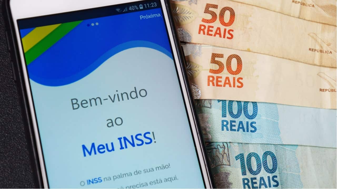 Celular com aplicativo Meu INSS ao lado de notas de 50 e 100 reais