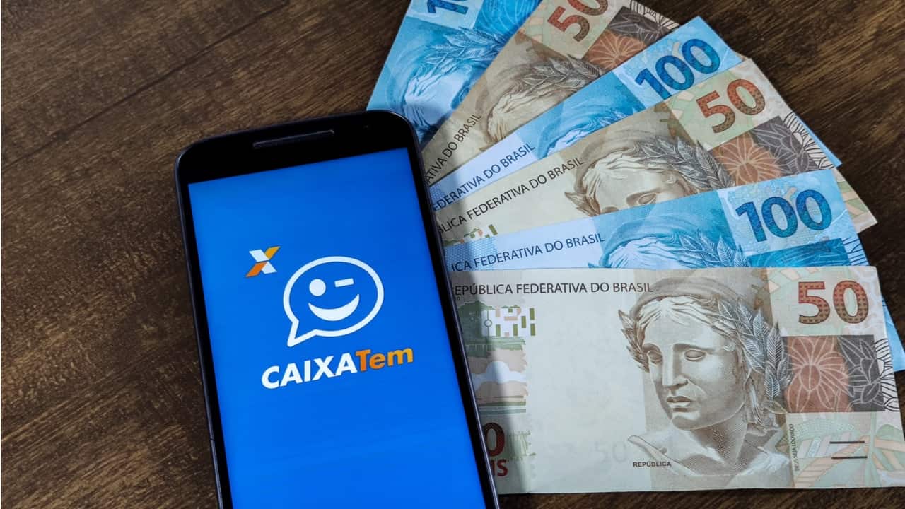 Celular posicionado sobre mesa, com o logo do aplicativo Caixa Tem na tela, com notas de 50 e 100 reais ao lado do aparelho