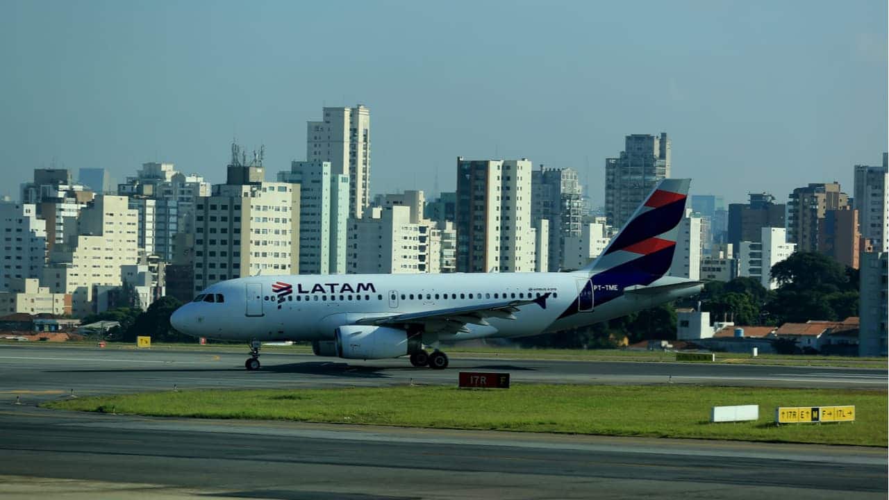 Imagem focada em um avião da companhia aérea LATAM. Ao fundo, prédios residenciais.