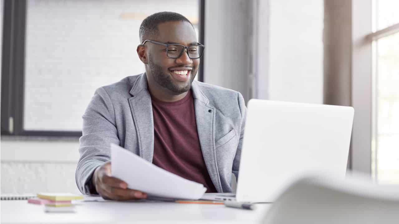 Imagem de jovem negro que usa óculos sorrindo enquanto olha para seu notebook