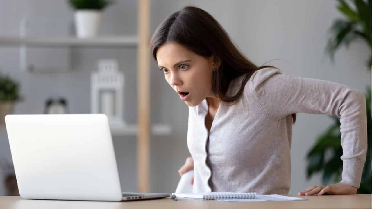 Mulher branca usando uma camisa branca olhando para o seu computador em estado de choque ao perceber que fez compra em site falso