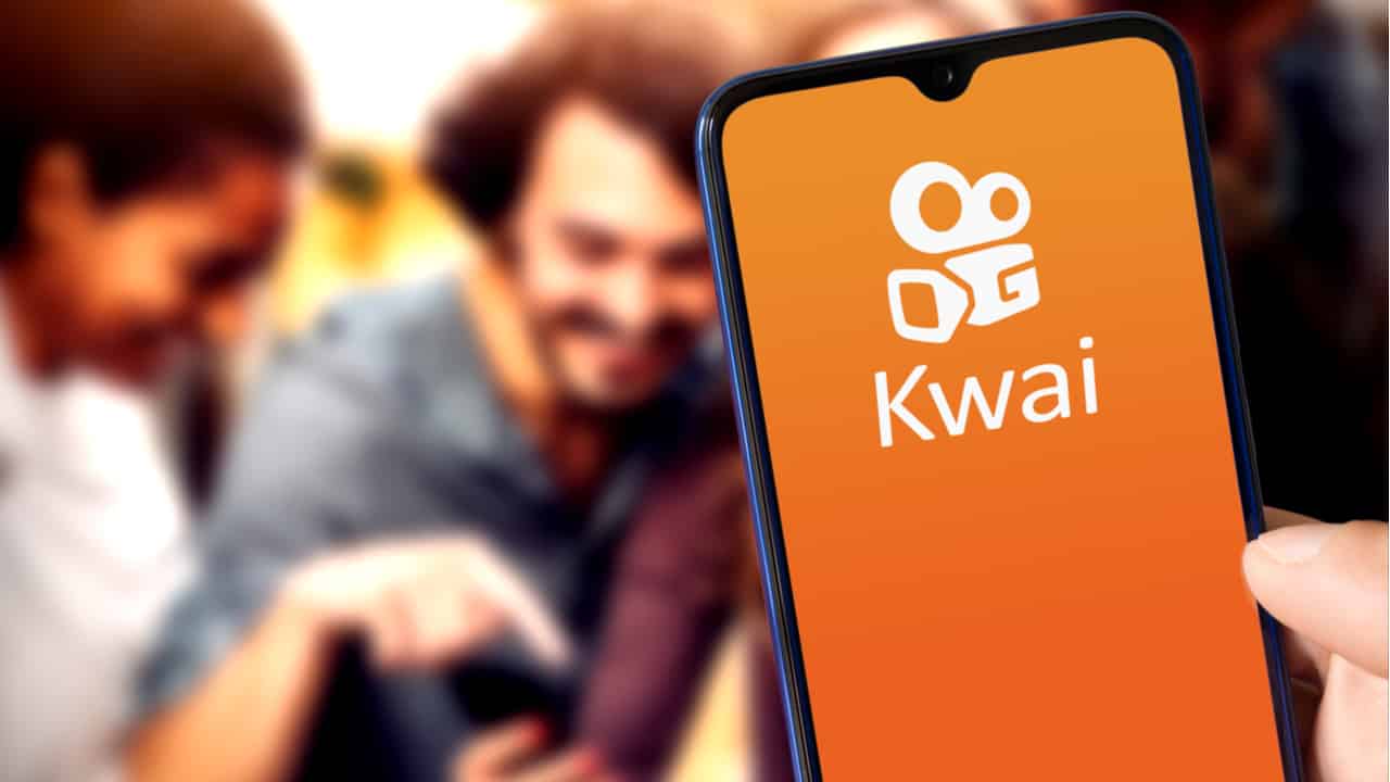 Mão segurando celular que mostra logo do Kwai e pessoas ao fundo