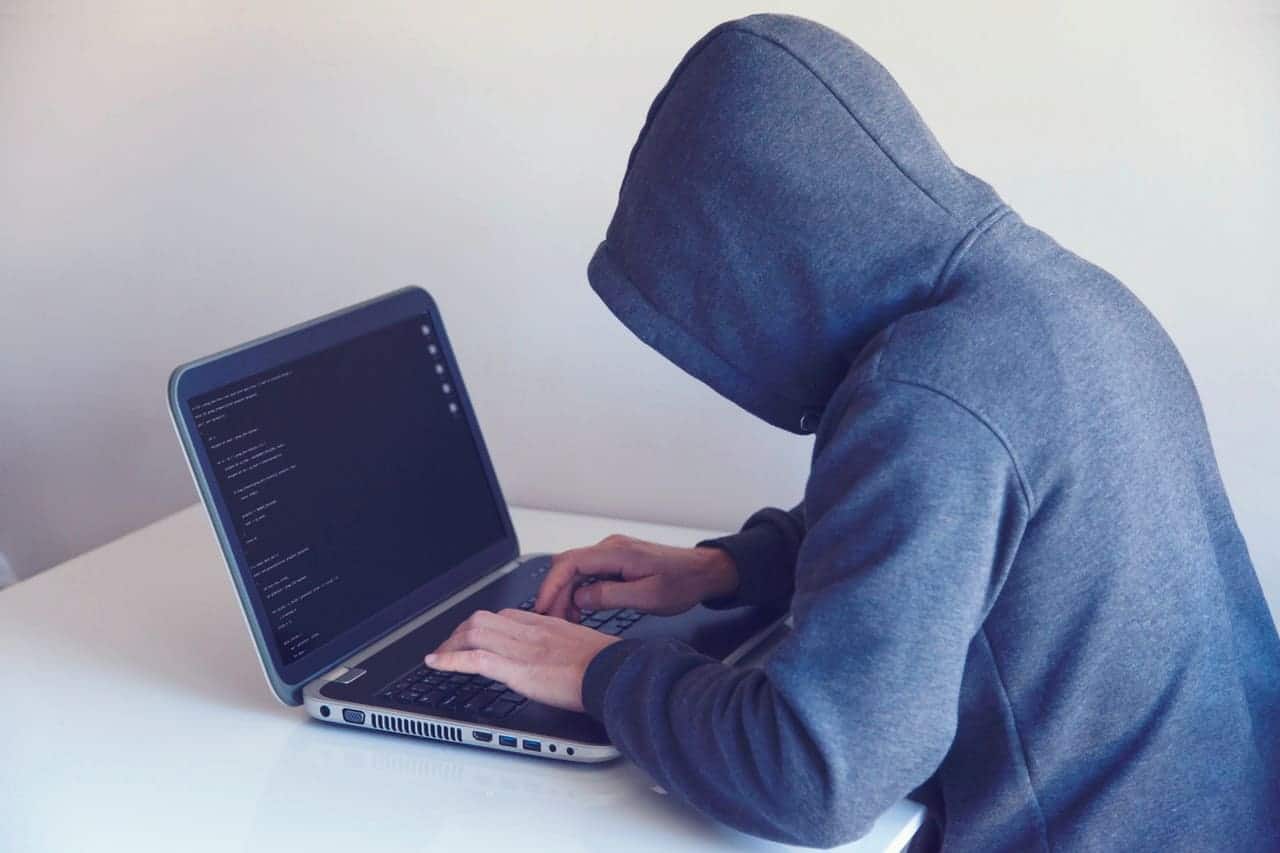 Homem sentado à mesa, vestindo um casaco azul com capuz, digitando em um notebook