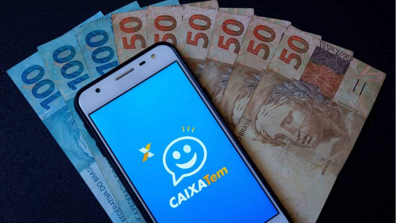 Aplicativo Caixa Tem sendo exibido em um celular com notas de 50 e 100 reais