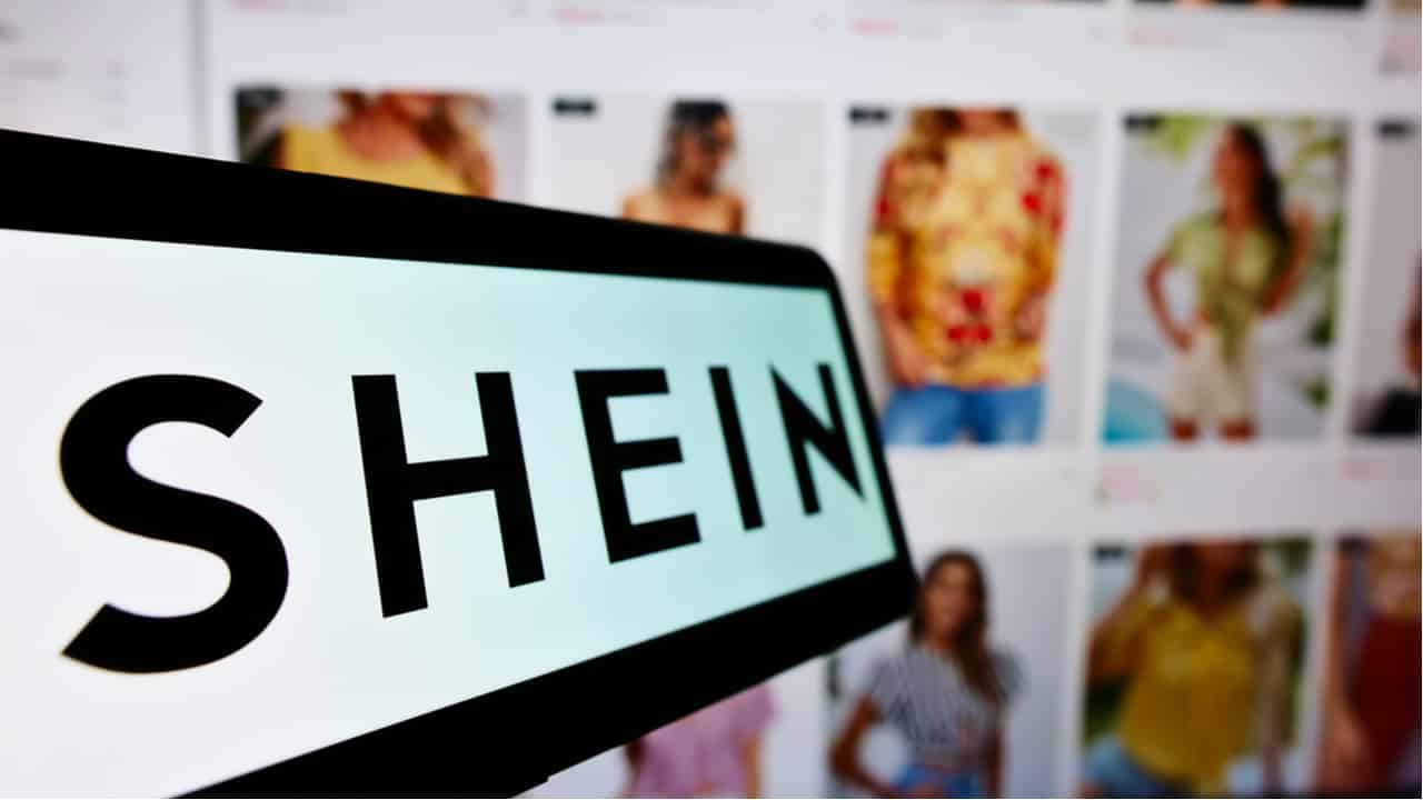 Logo da Shein, com uma página do site ao fundo, mostrando diferentes modelos com diferentes roupas.