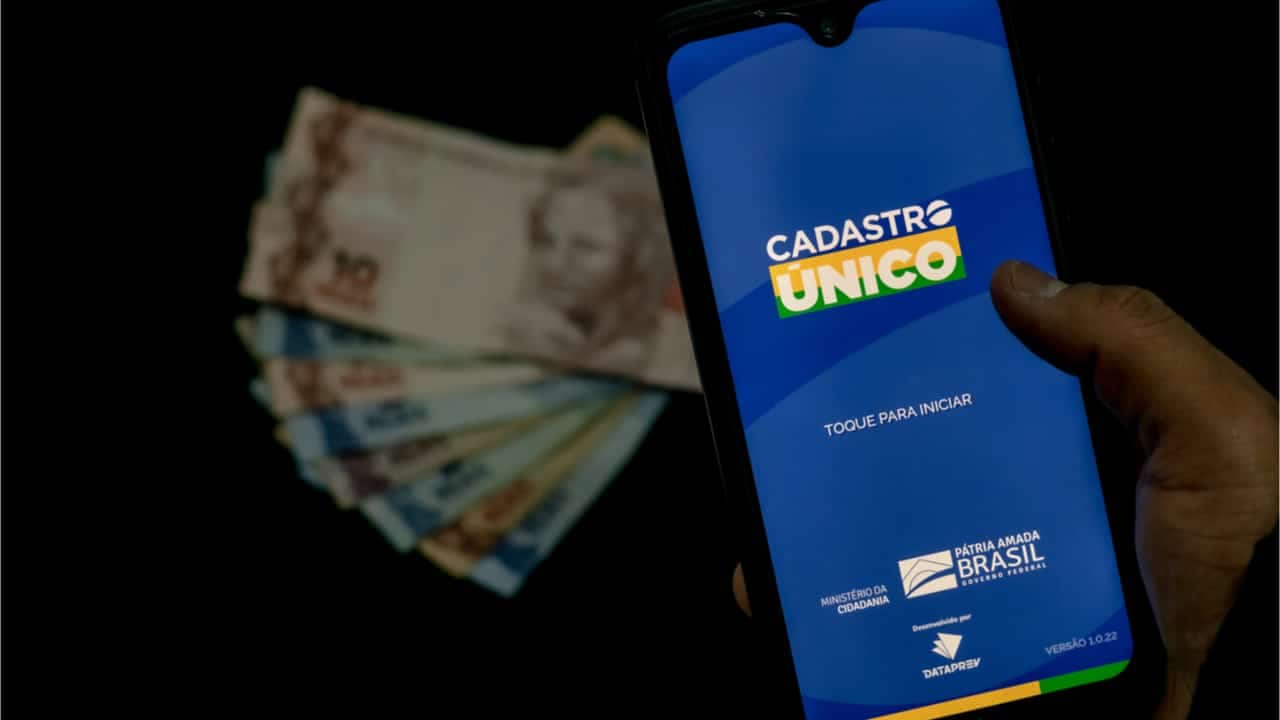 cédulas de Real em cima de uma mesa, sobreposta por um telefone mostrando o aplicativo do Cadastro Único - benefícios sociais