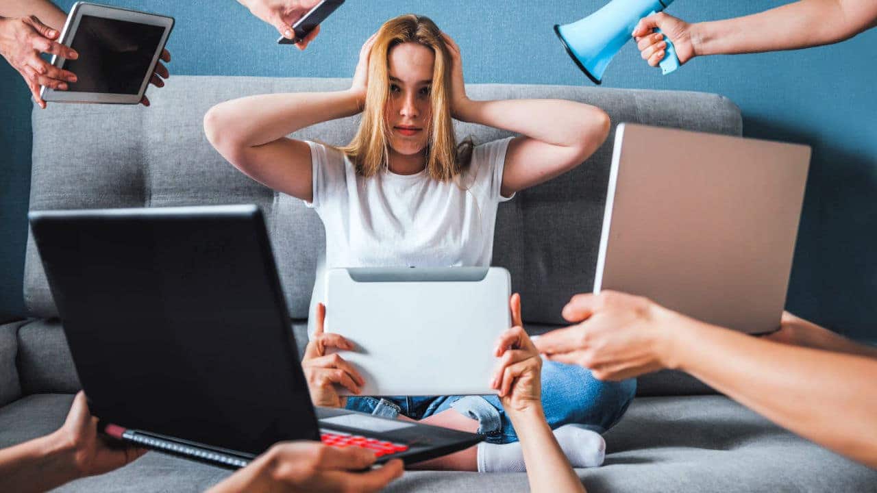 Mulher de ouvidos tapados ao ser bombardeada com informações na internet representadas por megafone, celular e tablet