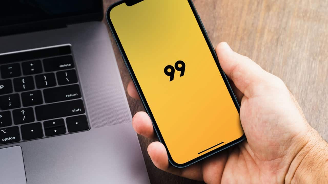 Imagem do aplicativo 99 em celular, com notebook ao fundo