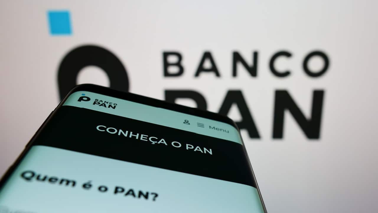 Em primeiro plano, tela de celular mostrando site do Banco Pan. Ao fundo, aparece desfocado o logotipo da instituição financeira.