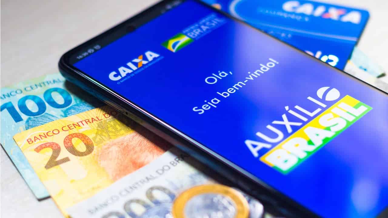 Celular com aplicativo do Auxílio Brasil ao lado de cédulas e moedas de real e um cartão da Caixa