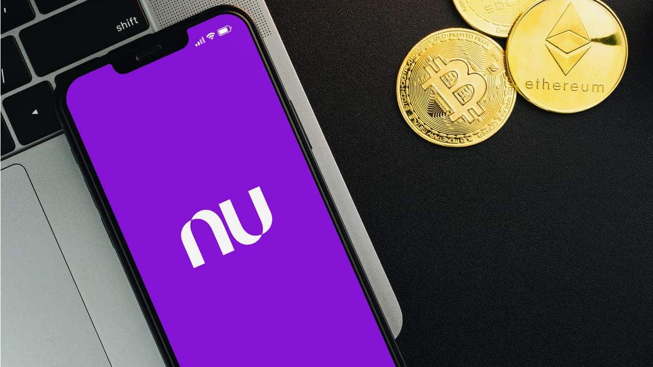 Celular com aplicativo do Nubank sobre teclado de computador. Ao lado, moedas com símbolos das criptomoedas Bitcoin e Ethereum