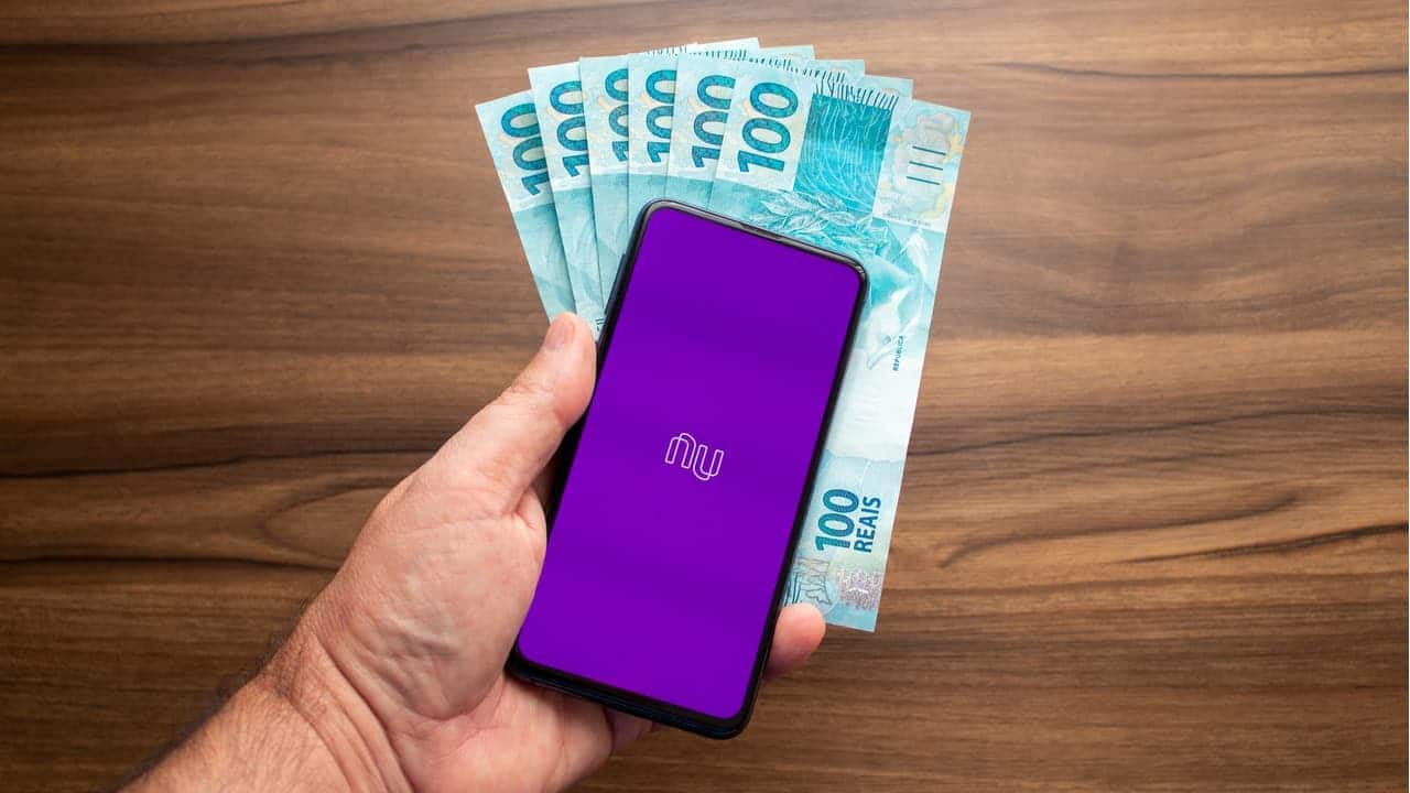 Mão segurando um smartphone com aplicativo do Nubank e notas de R$ 100 ao fundo