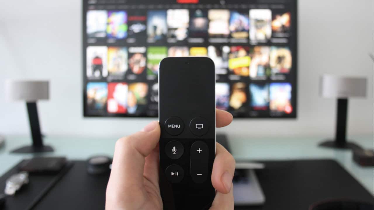 Mão segurando controle remoto e TV ao fundo mostrando catálogo de filmes ou séries