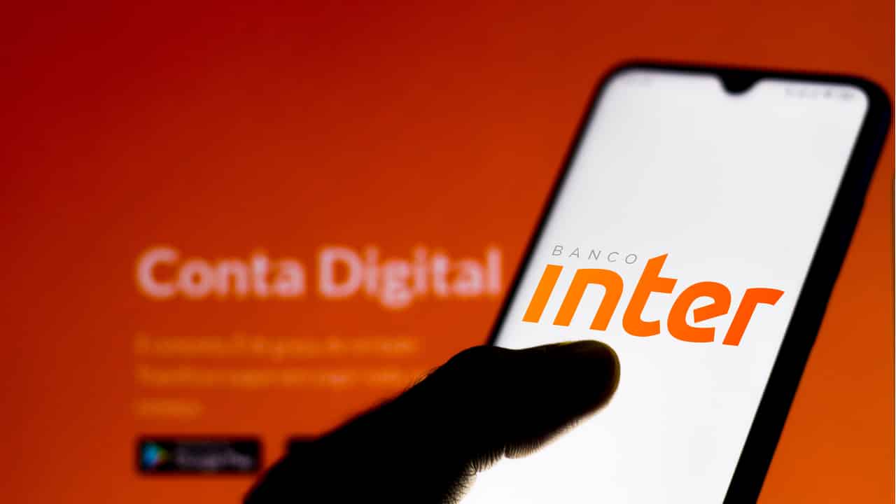 Sombra de uma mão segurando celular que mostra logo do banco Inter. Ao fundo, desfocada, uma tela que lembra o site do banco mostra as palavras "conta digital".