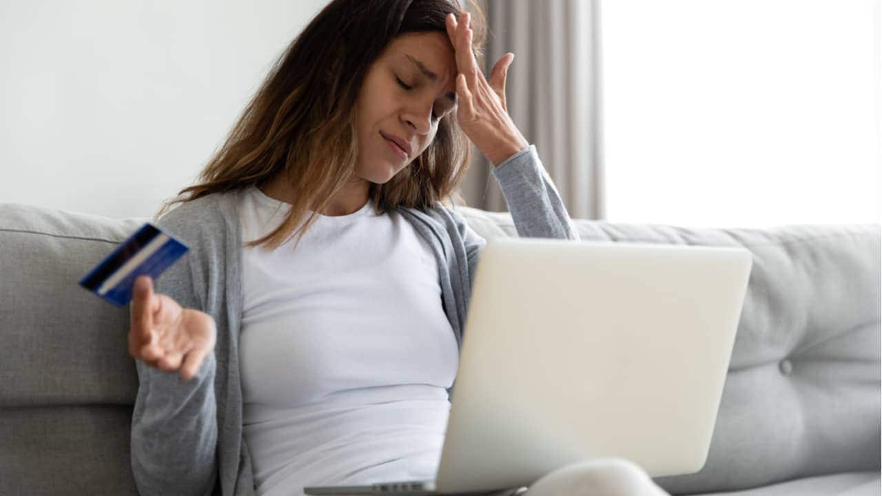 Mulher lamenta com a mão na testa, segurando cartão de crédito e com notebook no colo