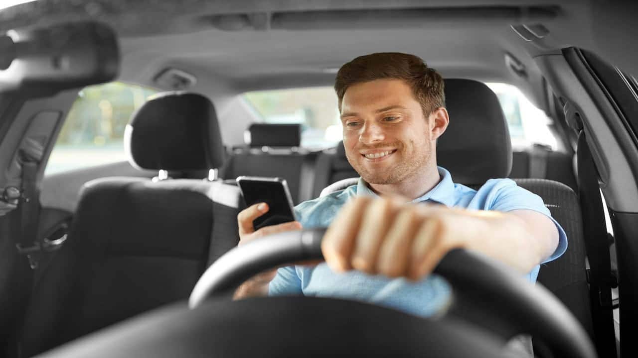 Motorista segurando volante de um carro e olhando para seu celular