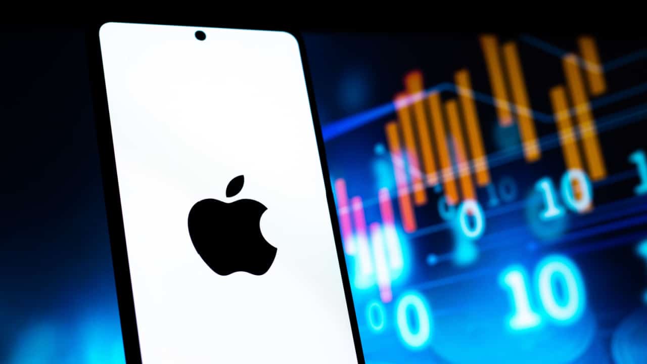 Celular mostrando logo de maçã da Apple. Fundo desfocado com gráficos e números coloridos.