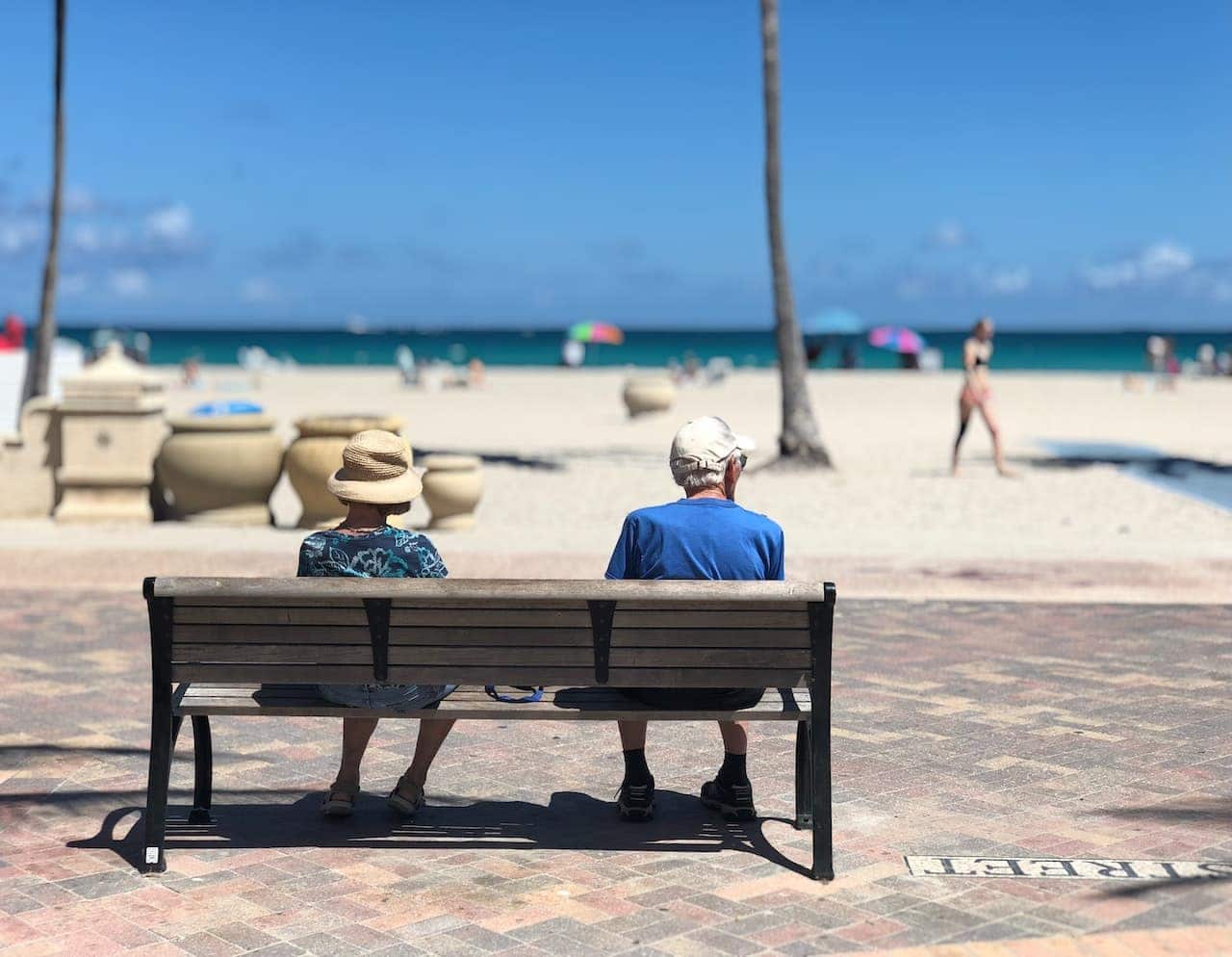 Fotografia em que um casal de senhores, sentado em um banco, observa a vista da praia.