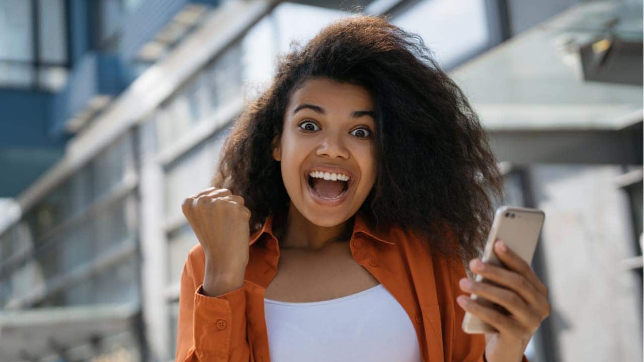 Mulher jovem olhando em direção à câmera e comemorando, com celular na mão