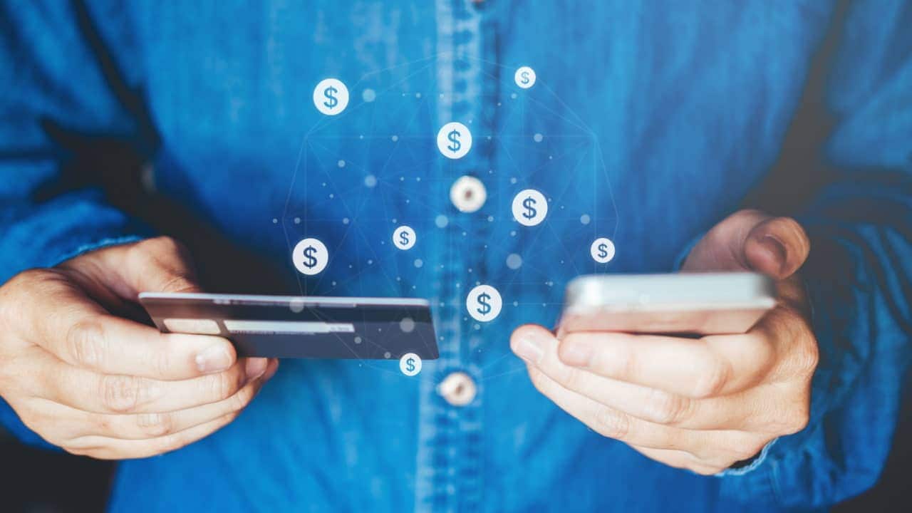 Pessoa segurando cartão de crédito com uma mão e celular ao lado, com a outra mão. Entre os dois objetos, cifrões de dinheiro flutuando.