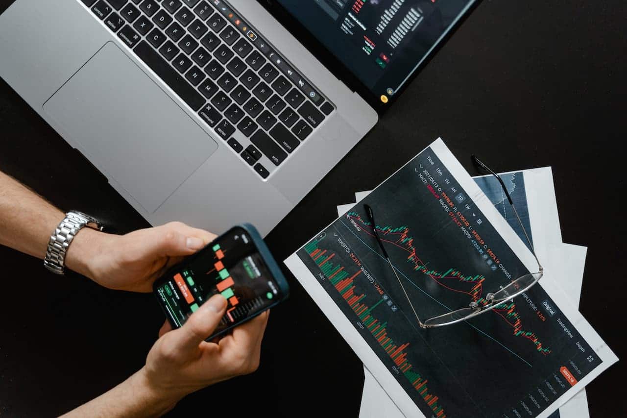 pessoa opera na bolsa de valores pelo celular, enquanto outras duas telas apresentam gráficos de mercado financeiro.
