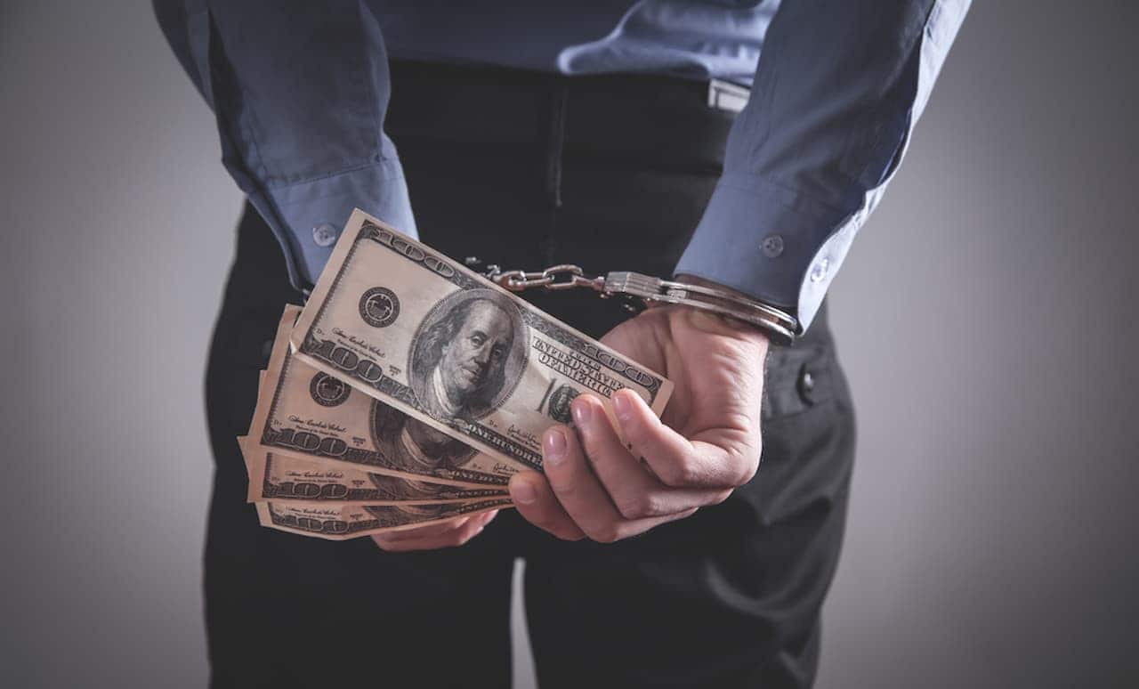Pessoa com mãos algemadas nas costas e segurando notas de dólar