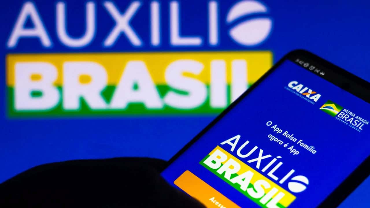 Celular mostrando tela inicial aplicativo Auxílio Brasil. Ao fundo, logo do programa.