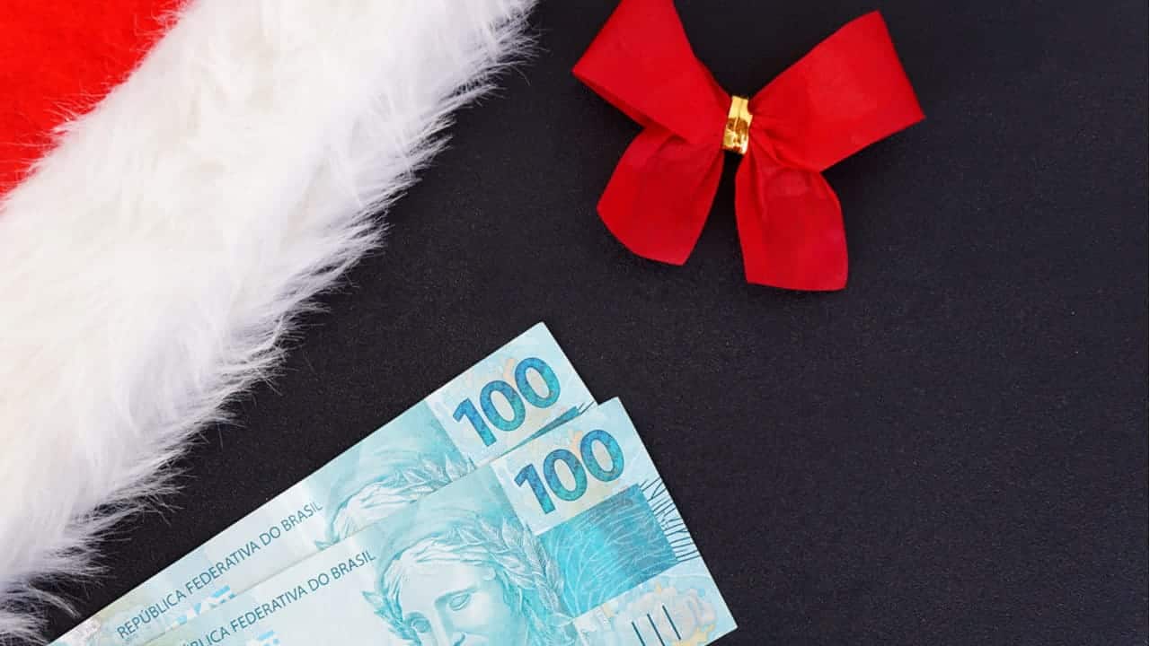 Notas de cem reais em fundo com parte do gorro do Papai Noel e laço natalino.