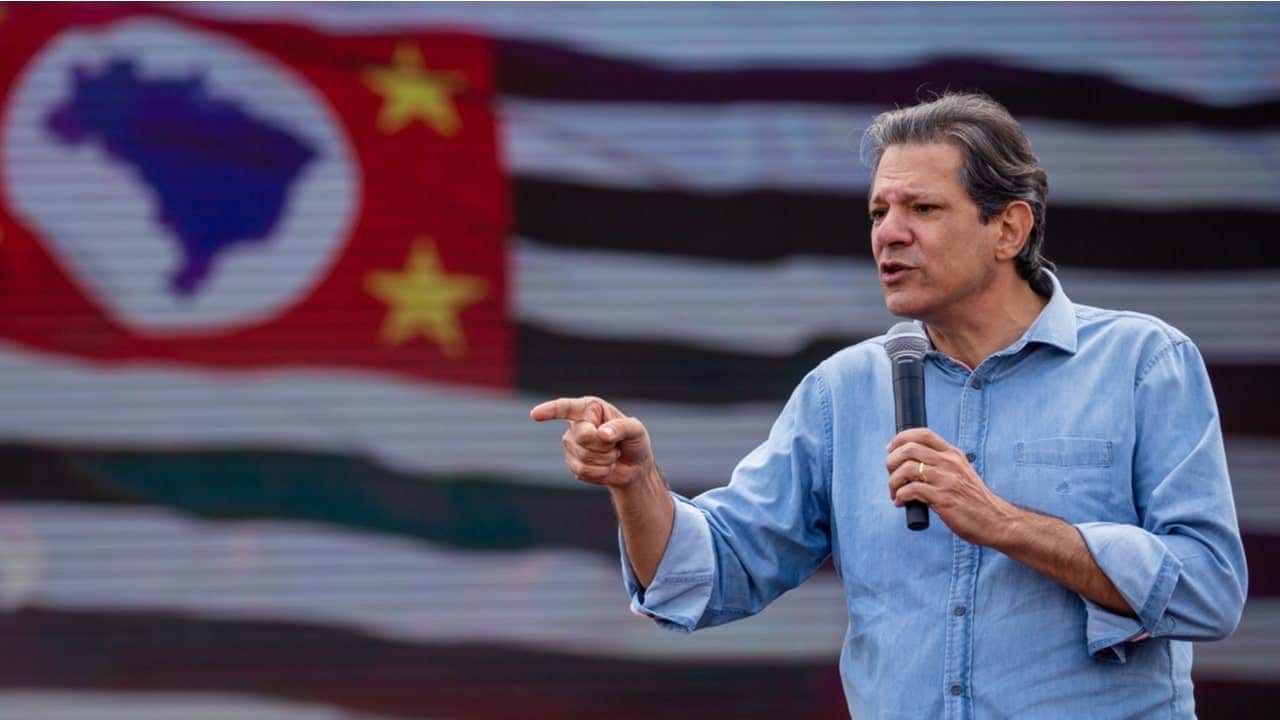 Fernando Haddad discursando com microfone e bandeira de São Paulo desfocada ao fundo