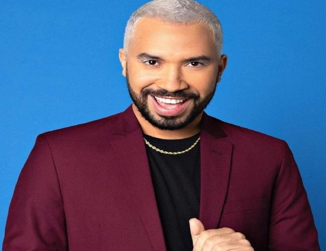 Foto em que GIl do Vigor, de cabelo descolorido, aparece sorrindo vestindo uma blusa preta, um paletó vinho e utilizando um colar prata.