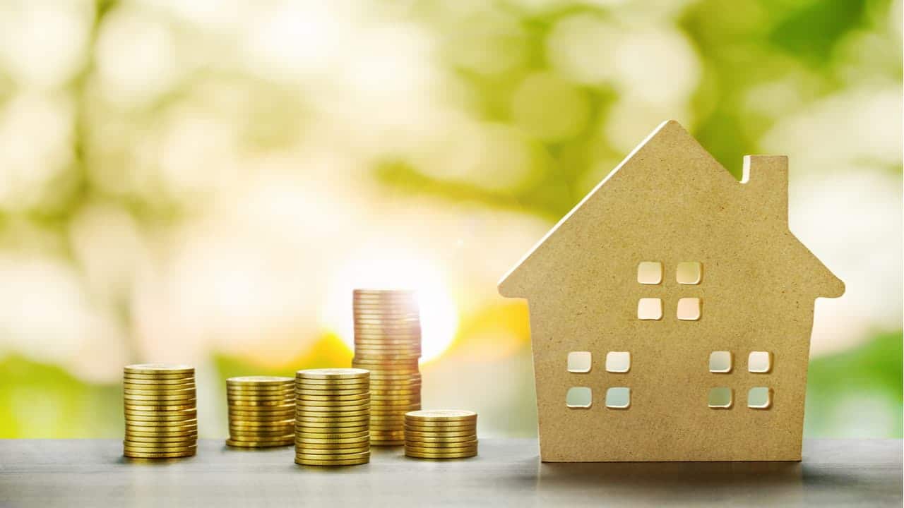 Quatro pilhas de moedas ao lado de miniatura com formato de casa. Ao fundo, vegetação desfocada