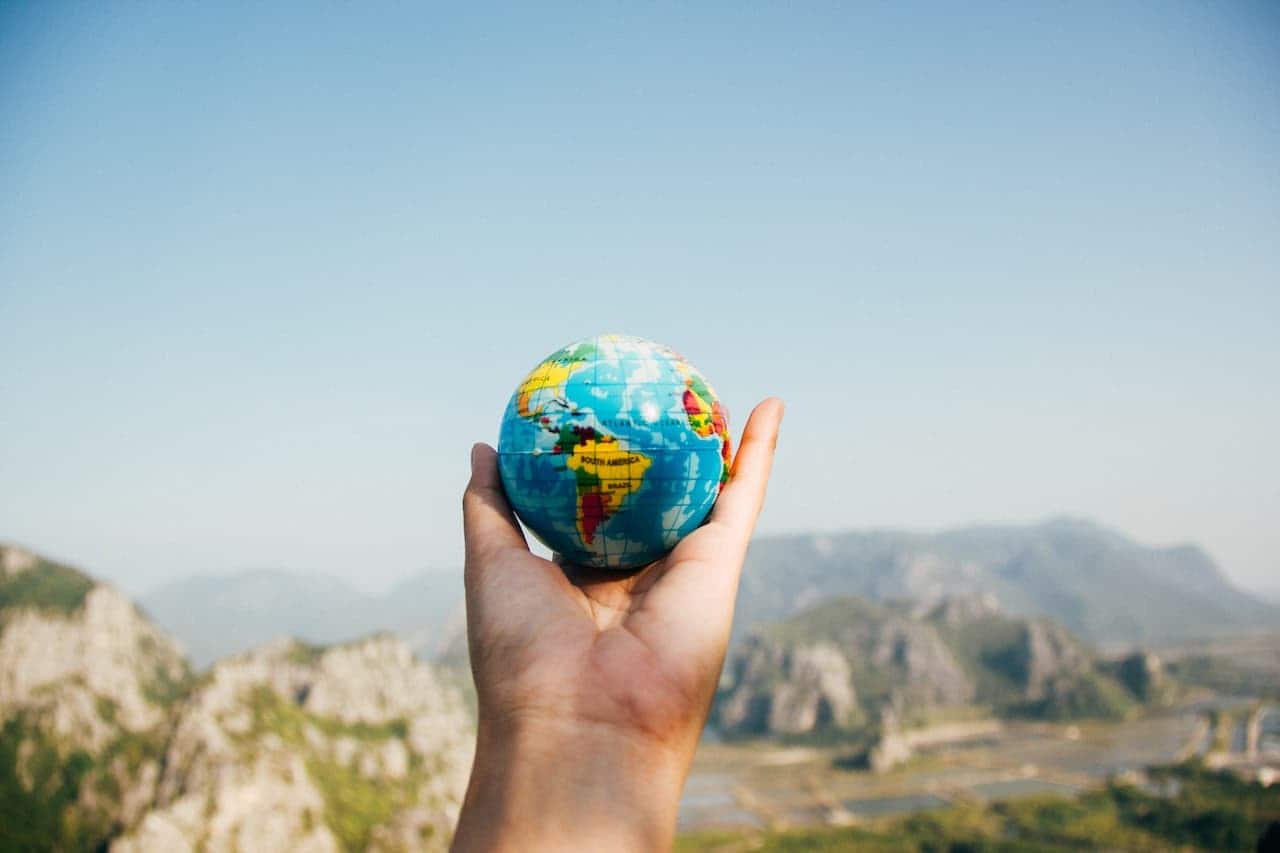 Mão de uma pessoa segurando miniatura do globo terrestre. Ao fundo, paisagem montanhosa desfocada.