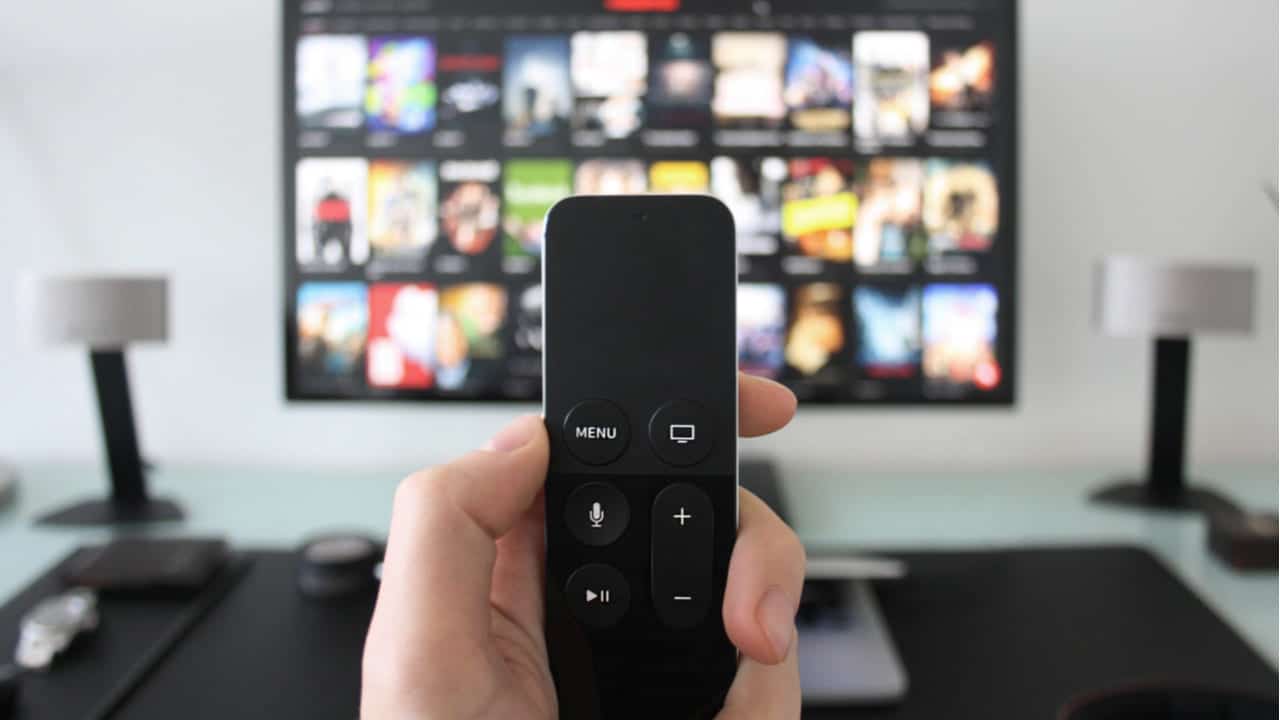 Mão segurando controle remoto e TV ao fundo mostrando catálogo de filmes ou séries