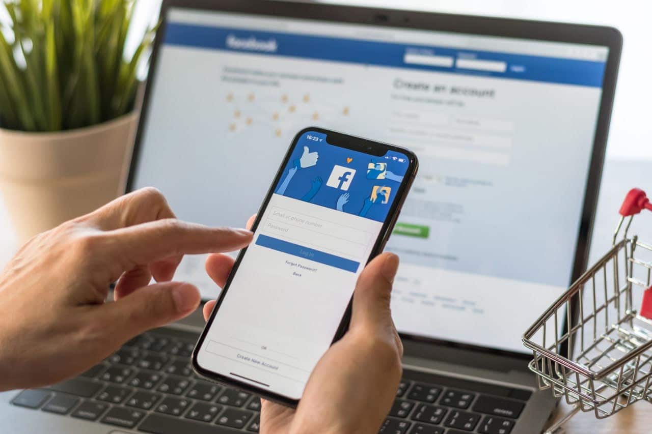 Mãos de uma pessoa usando celular com aplicativo do Facebook. Ao fundo, tela de notebook com o site do Facebook aberto.