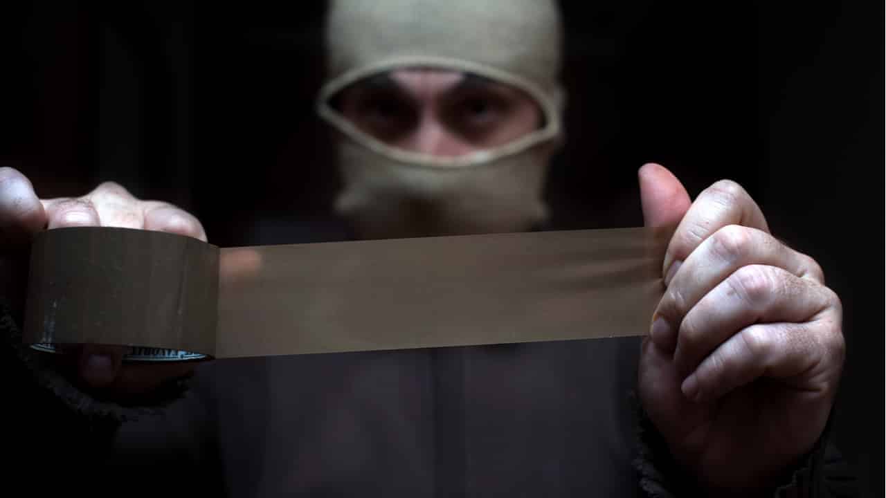 Sequestrador encapuzado segurando fita adesiva. Fundo preto