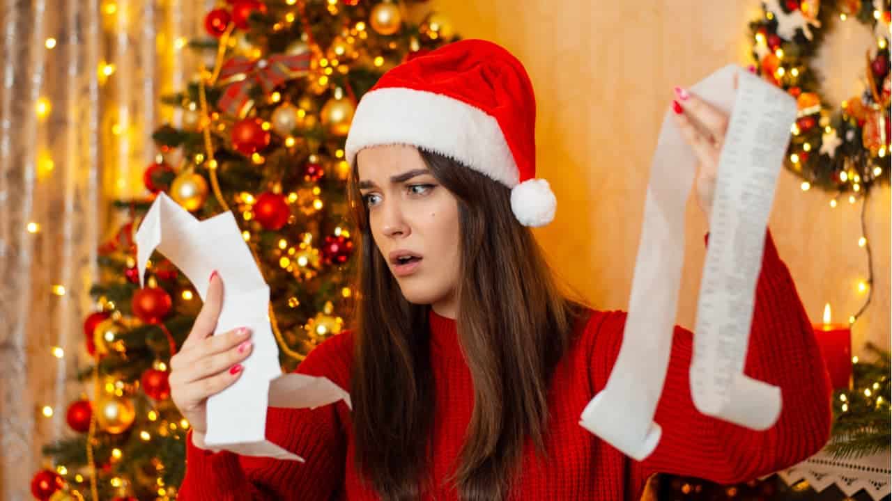 Moça usando touca de Papai Noel e com decoração de natal no fundo chocada com o valor que está na nota fiscal de compras, muito grande, que segura
