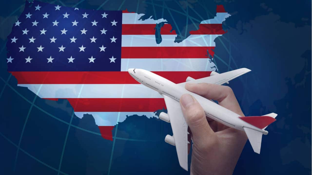 Mão segurando miniatura de avião sobre mapa dos Estados Unidos preenchido com a bandeira do país