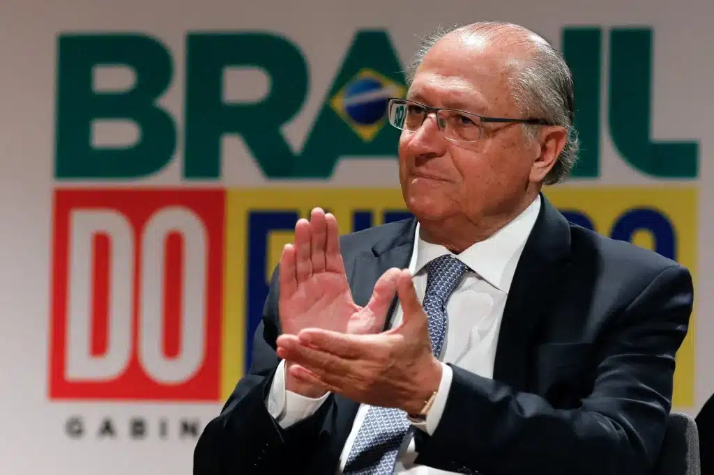 Geraldo Alckmin batendo palma, no fundo, o logo do novo governo