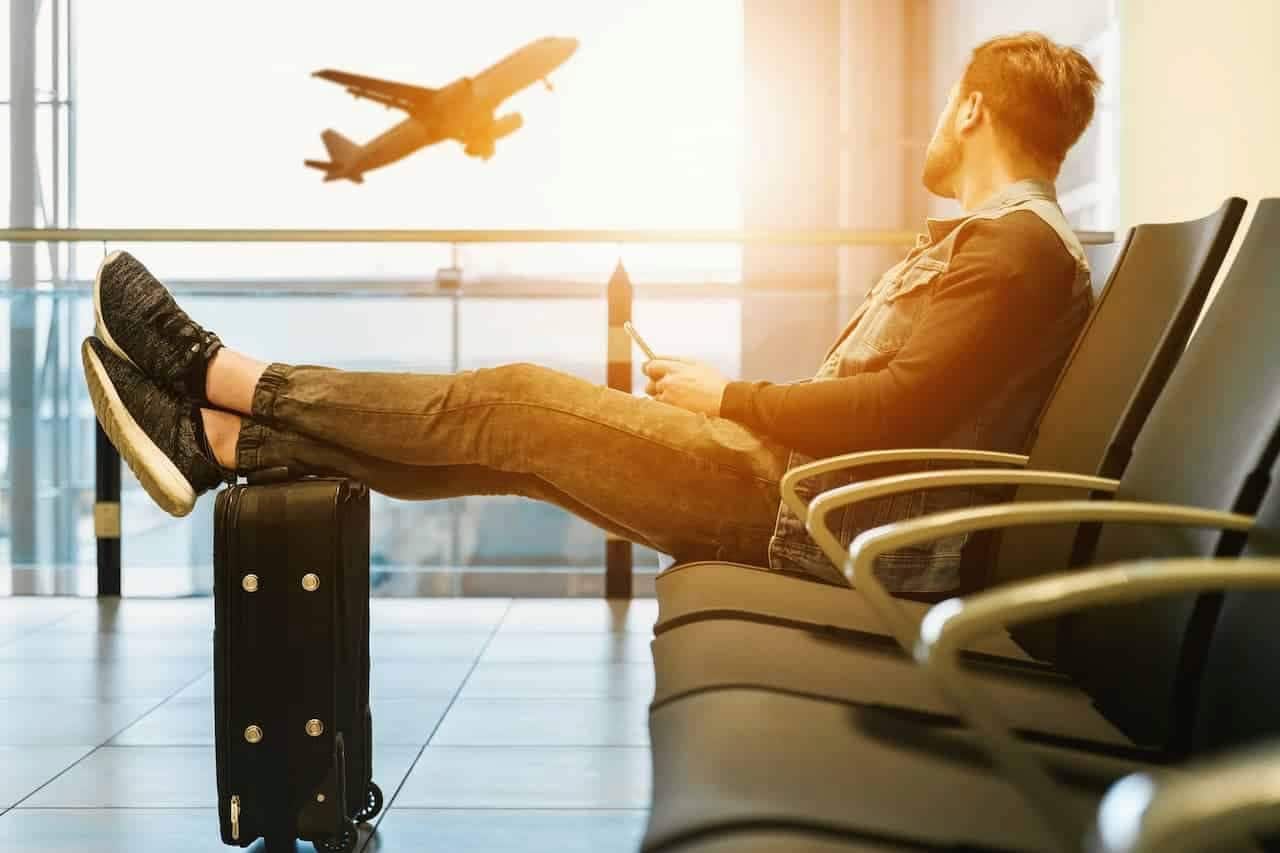 Um homem sentado em uma cadeira dentro do aeroporto olhando para um avião decolando